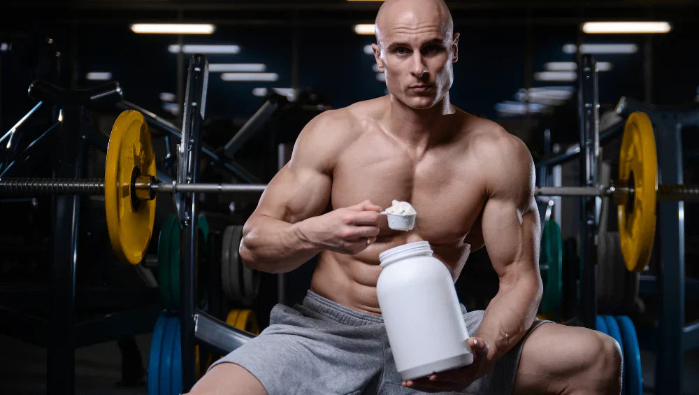 Homme sec et musclé torse nu assis sur un banc de developper couché en train de tenir un pot de whey avec une scoop pleine de protéine dans l'autre main