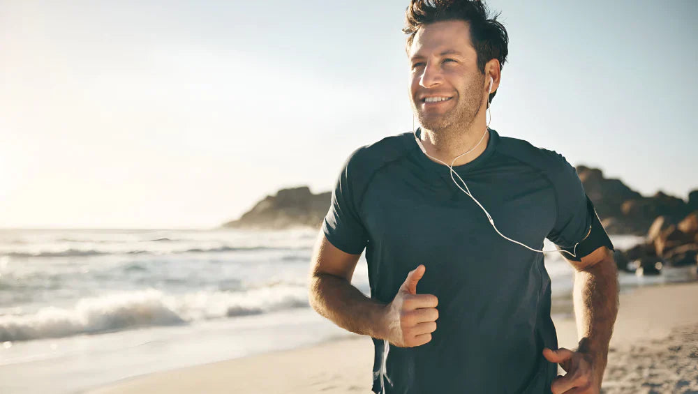 Homme souriant en train de faire un footing avec des écouteurs le long d'une plage avec en arrière plan une vue sur la mer