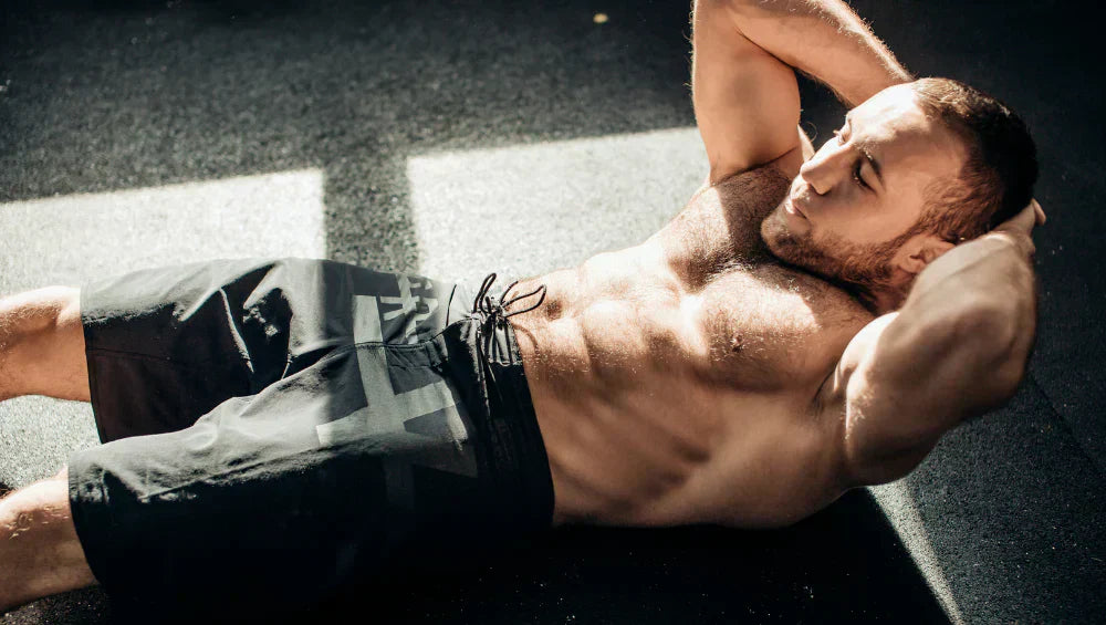 Homme musclé en train de faire une série d'abdos crunch dans une salle de sport laissant passer des rayons de soleil