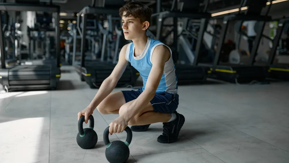 Adolescent accroupi dans une salle de sport tenant dans chacune des mains un kettlebell