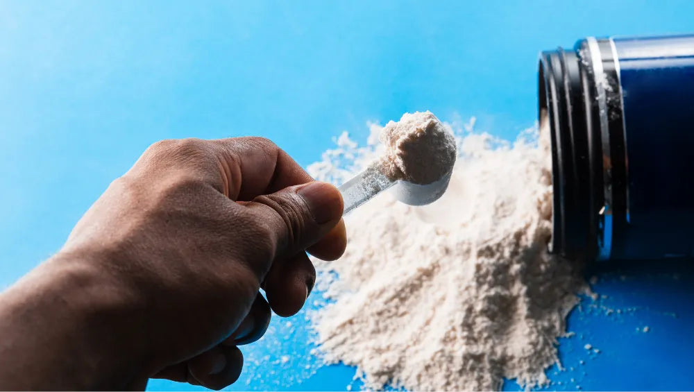 Une main tenant du bout des doigts une scoop contenant de la poudre avec pour fond un pot renversé et de la poudre blanche cassé qui en sort