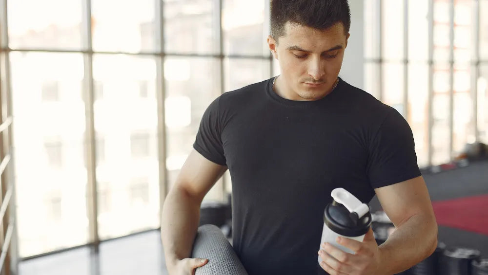 Homme en T-Shirt noir en train de regarder un shaker de protéine (whey) dans une salle de sport.