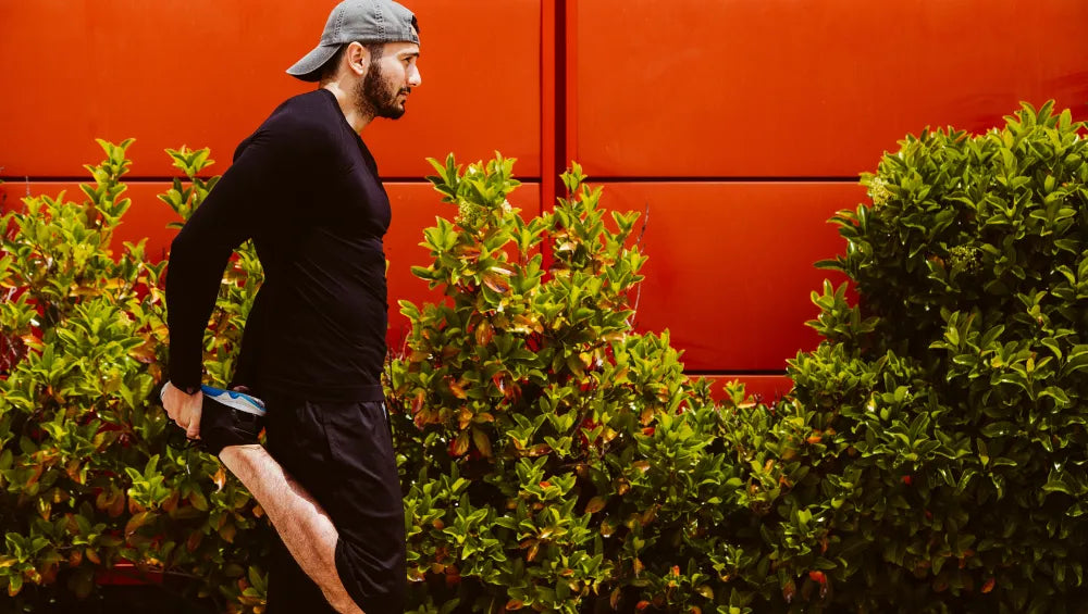 Homme en t-shirt manche longue, short noir et casquette grise en train de s'échauffer en vue d'un footing sur un fond rouge et des buissons en arrière plan.