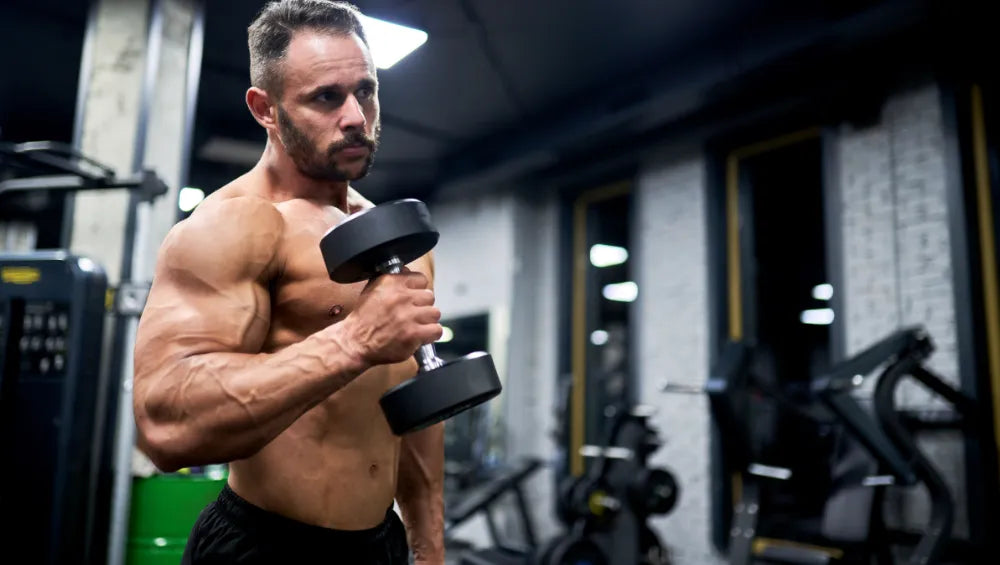 Homme musclé torse nu en train d'effectuer une série de curl biceps en prise marteau avec des haltères dans une salle de musculation.