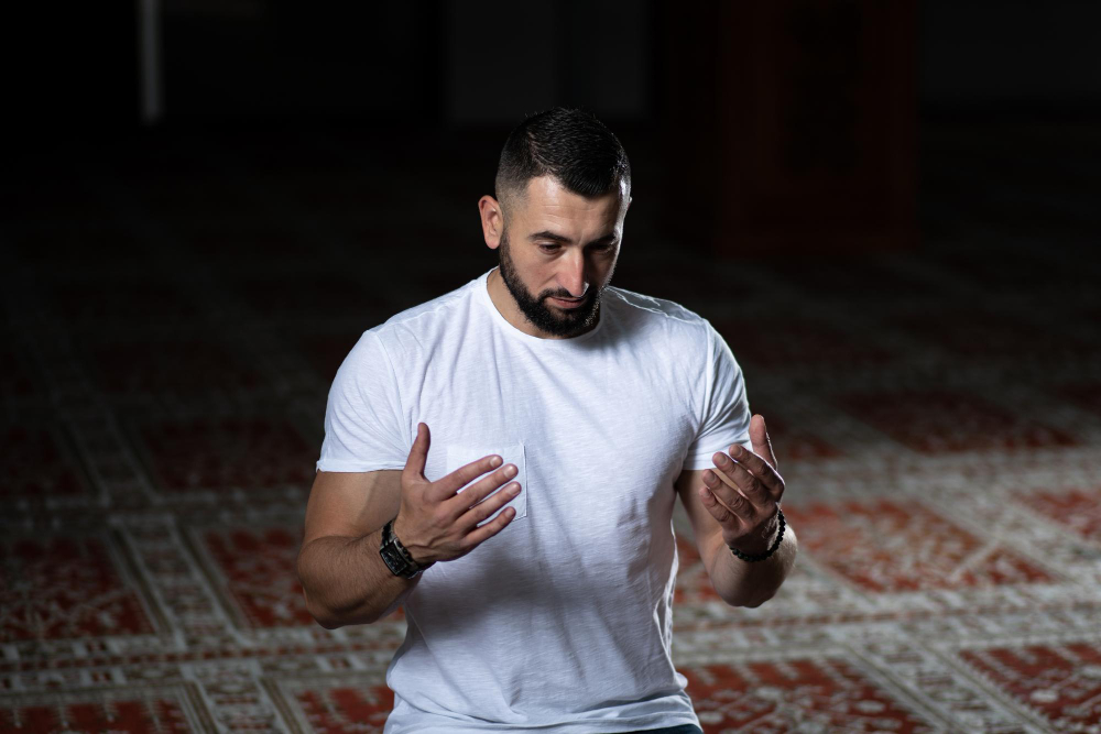 un jeune musulman en tshrt blanc agenouillé sur un tapis en train de prier 
